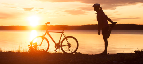Discover Maine's coastline on a bike. A woman biker takes a stretch break, watching the sun set over a placid lake.
